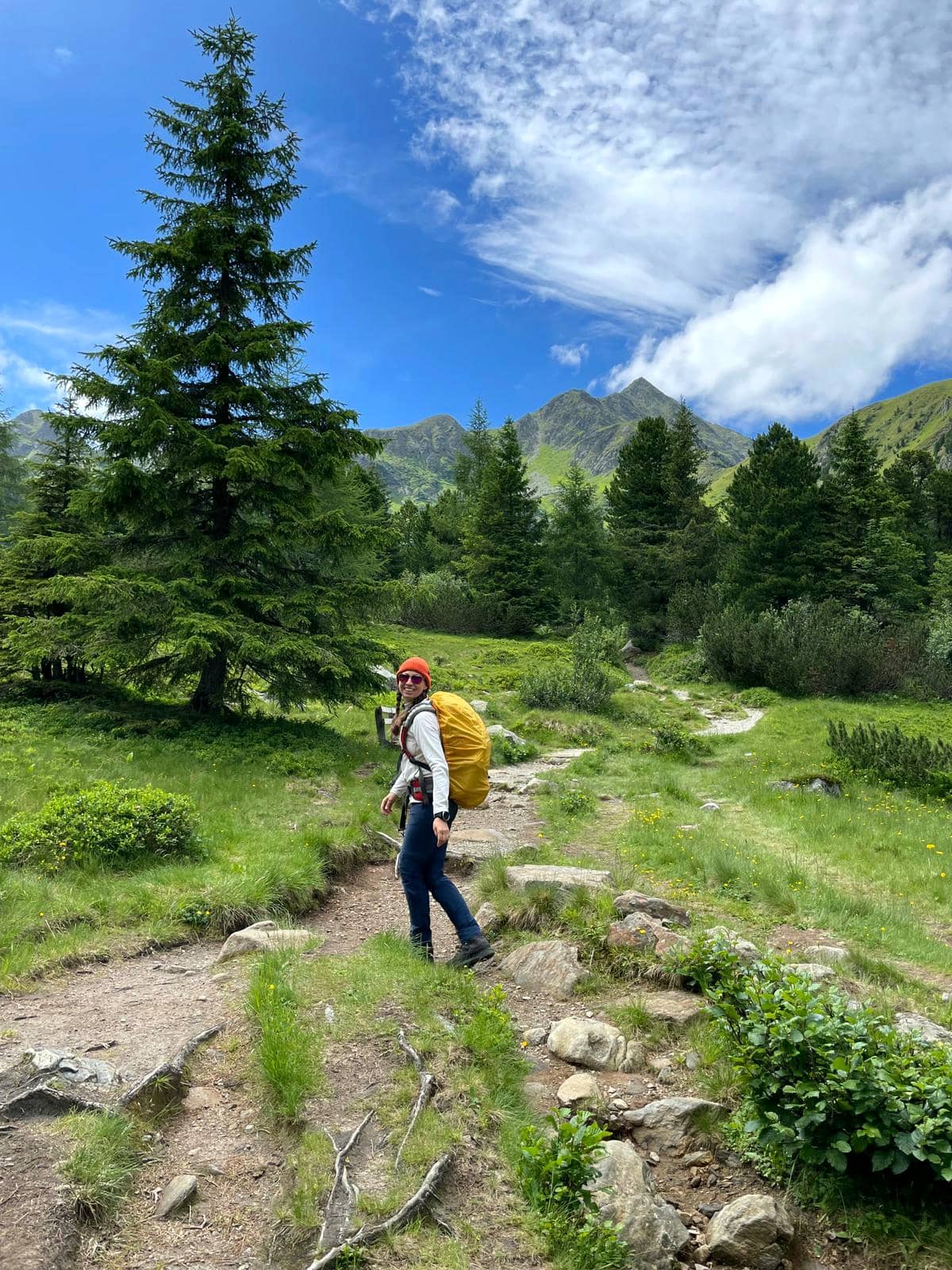 Frau in Wander-Outfit mit gelben Rucksack in Berglandschaft, blauer Himmel, Gras, grüne Bäume